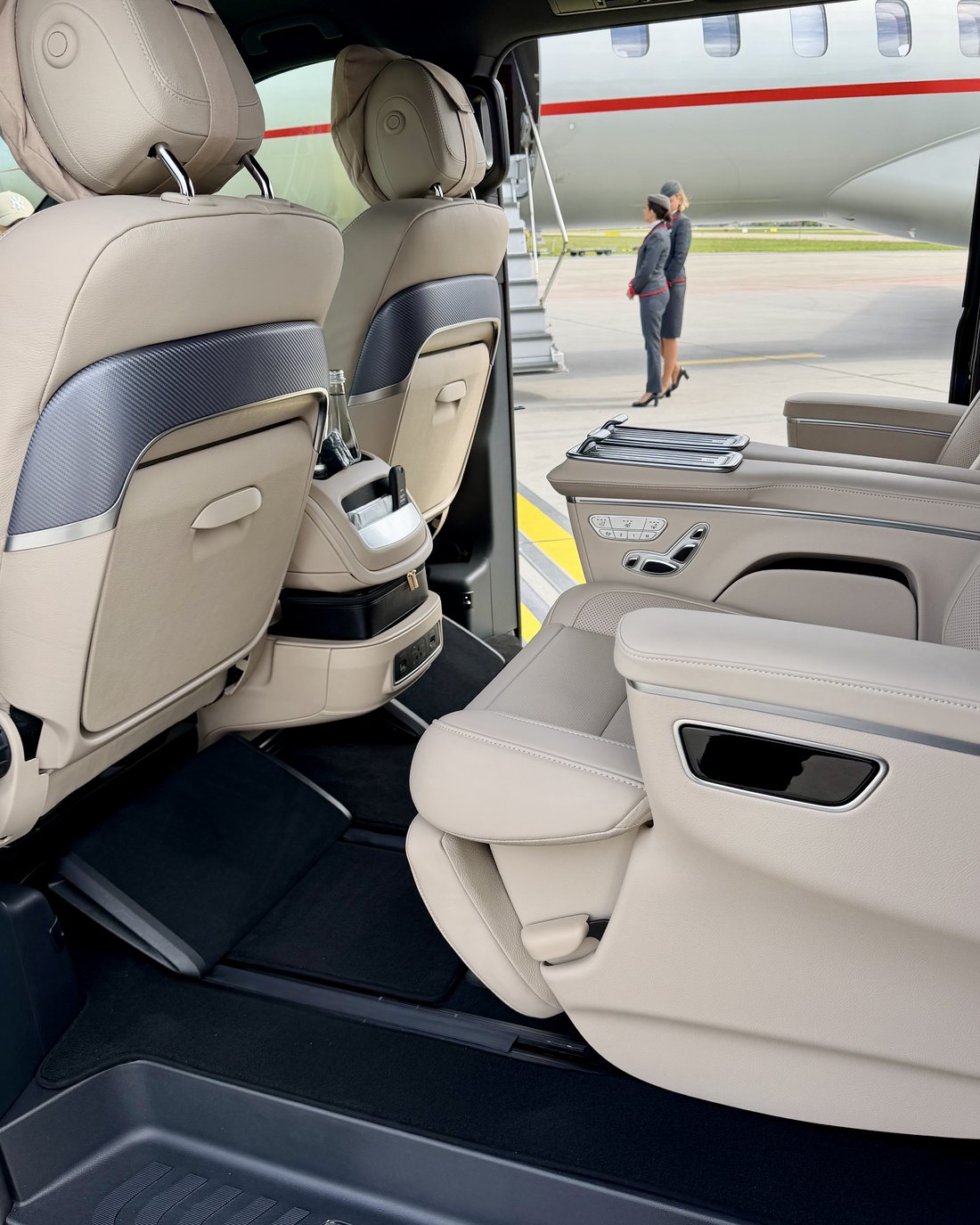 The view out of a Mercedes V-Class First Class through its open side door: cream leather seats in the foreground, and beyond them a private jet with two crew waiting at the airstair